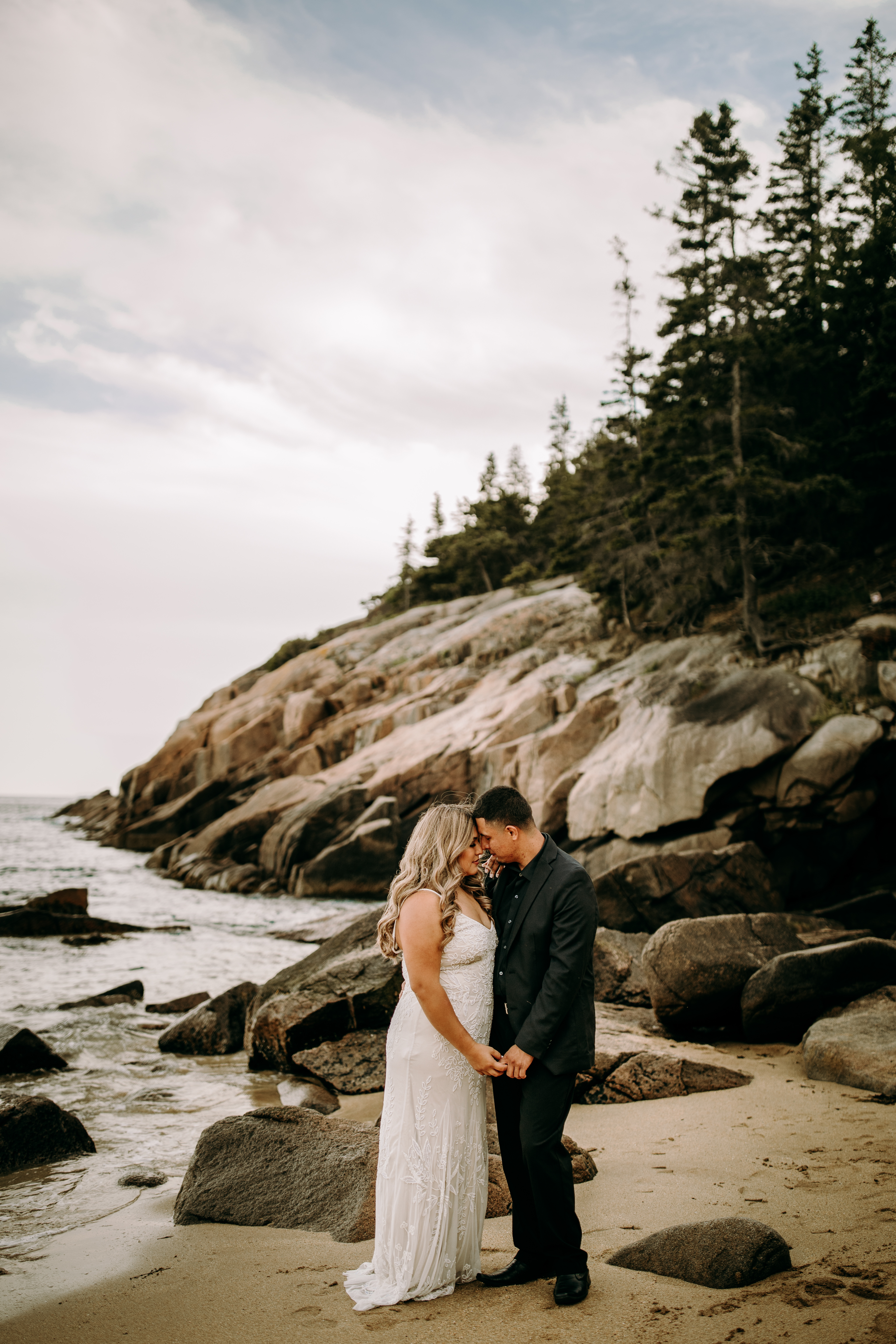 Couple facing each other pose at Acadia National Park in wedding dress and suit at Sand Beach with ocean in the background on the coast of Maine nature elopement intimate and documentary style wedding photography candid authentic 