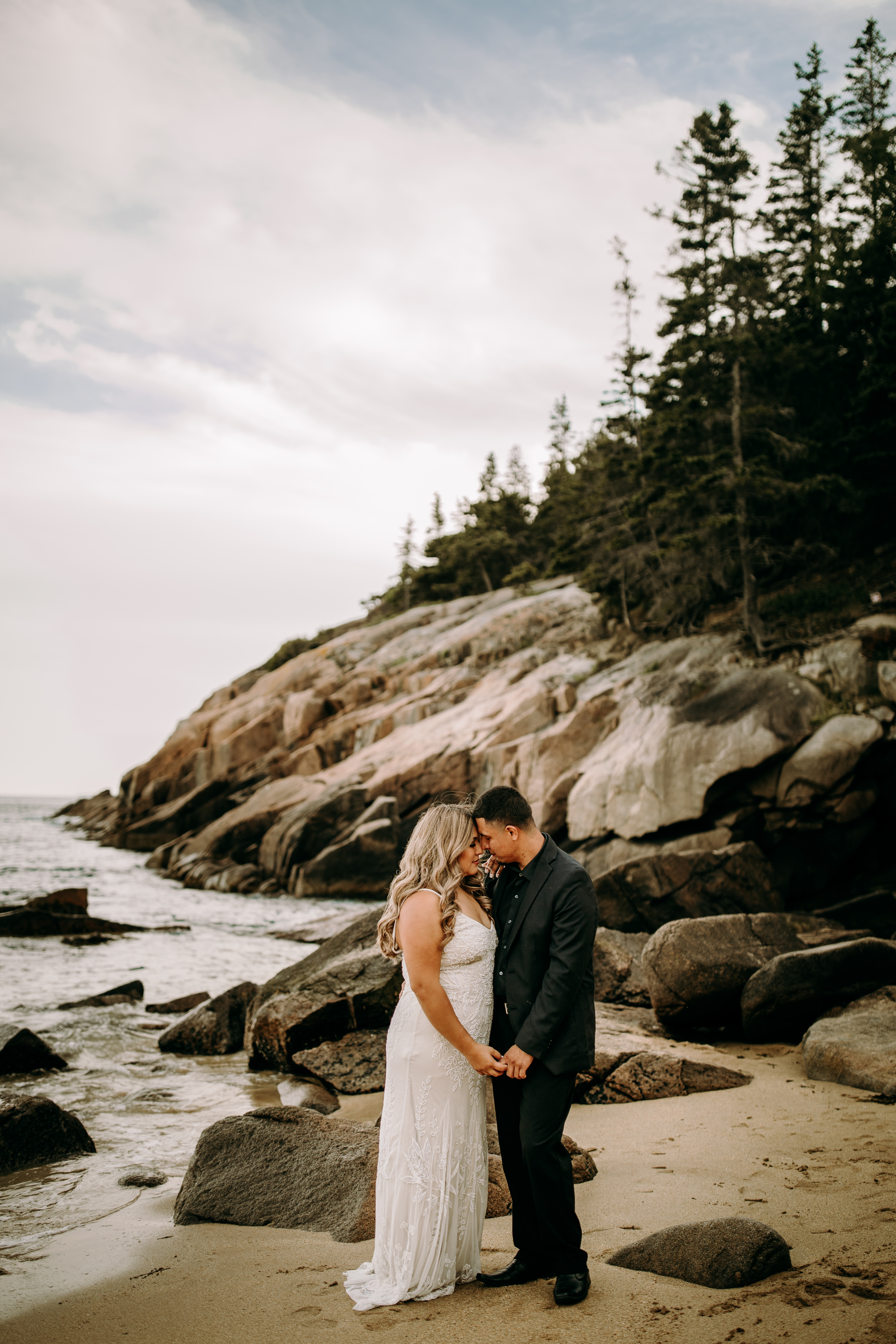Couple facing each other pose at Acadia National Park in wedding dress and suit at Sand Beach with ocean in the background on the coast of Maine nature elopement intimate and documentary style wedding photography candid authentic