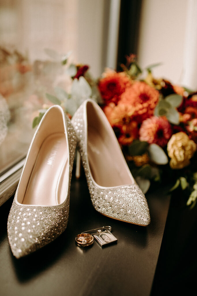 Wedding shoes and rings detail photo in Press Hotel Portland Maine with orange red and yellow bouquet in the background