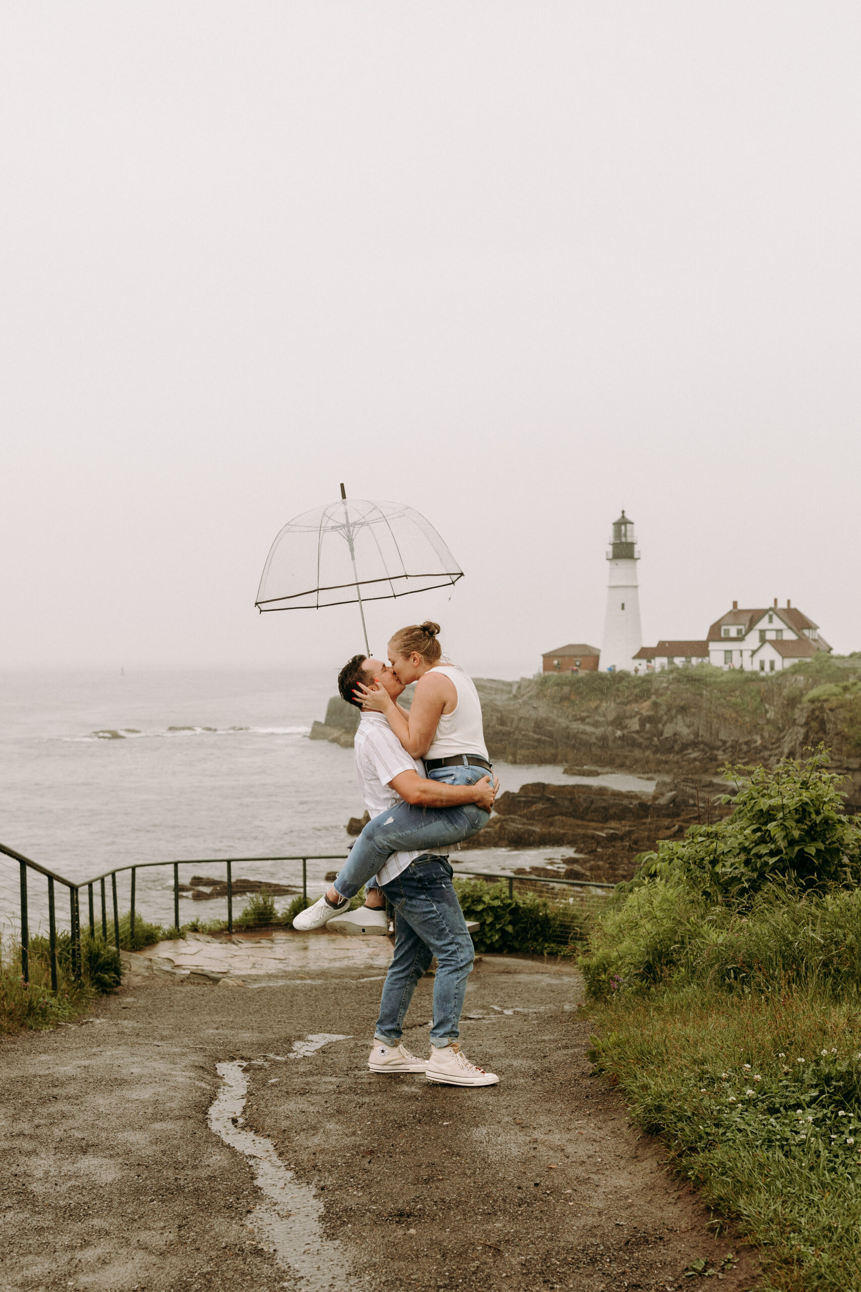An affectionate couple sharing a tender kiss in front of Portland Head Light, Maine. The iconic lighthouse stands tall in the background, while the couple's silhouettes are framed against the overcast sky. Their love shines brightly amidst the misty and romantic ambiance of the rainy day.