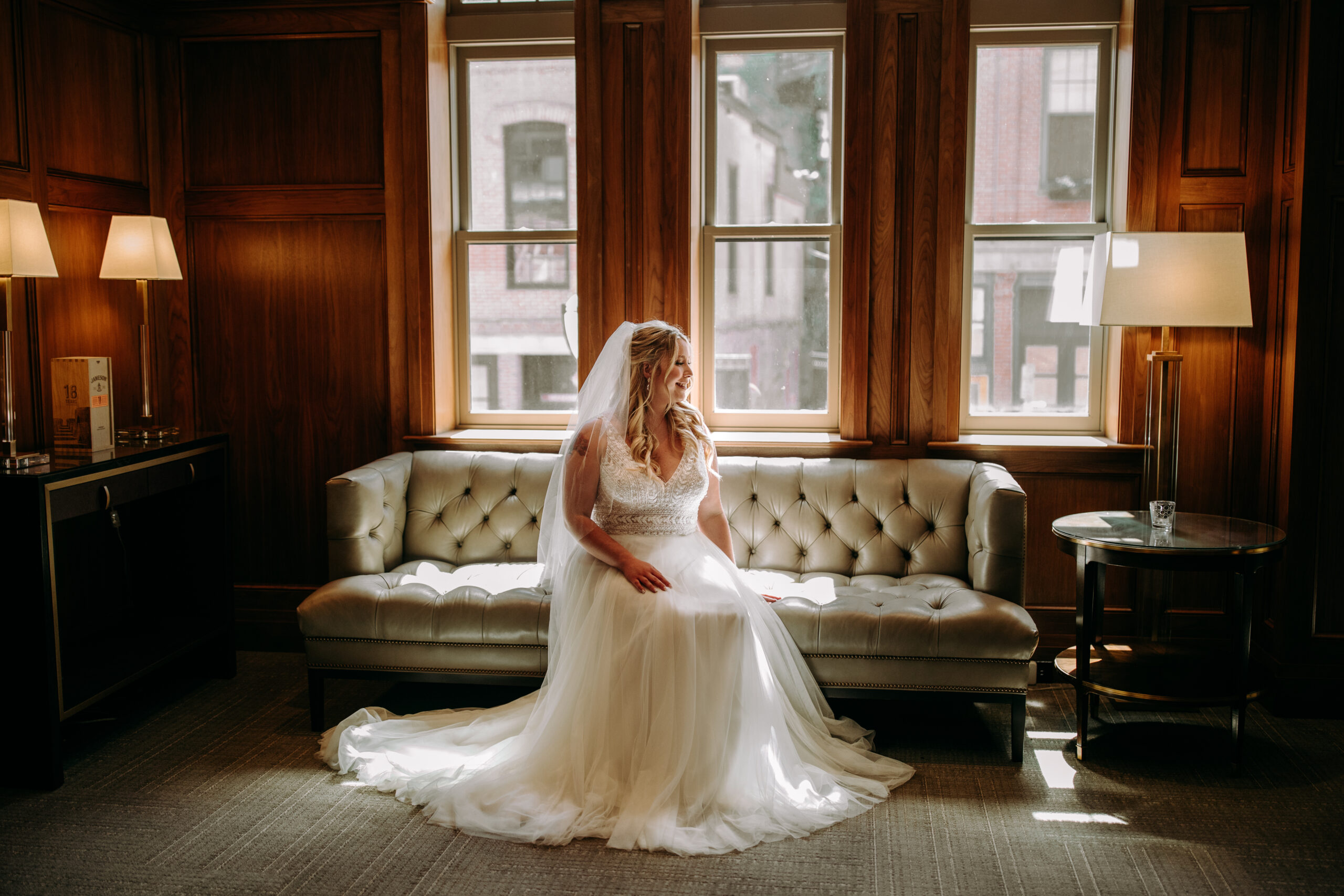 Blonde bride with her wedding dress and veil sitting on a tufted couch with natural light pouring in through the windows to backlight her while she waits for her wedding to start at the Portland Ocean Gateway in Portland Maine 