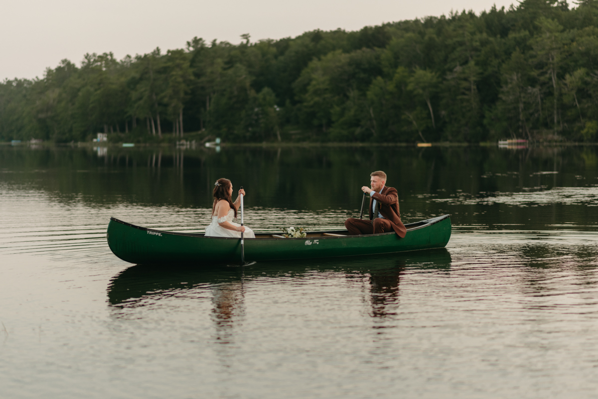 bride and groom in a canoe on their wedding day paddling in the middle of a serene pond surrounded by pine trees just after sunset blue hour