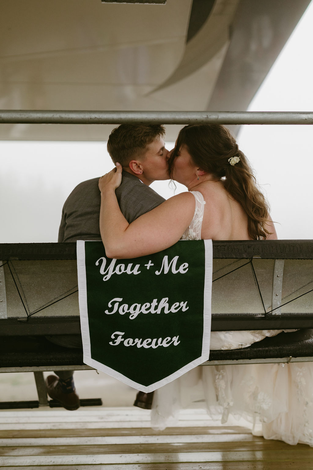 Brides, LGBTQ couple kissing with wedding banner that reads "you + me together forever" while sitting on a ski lift at Saddle Back Mountain in Rangeley Maine 