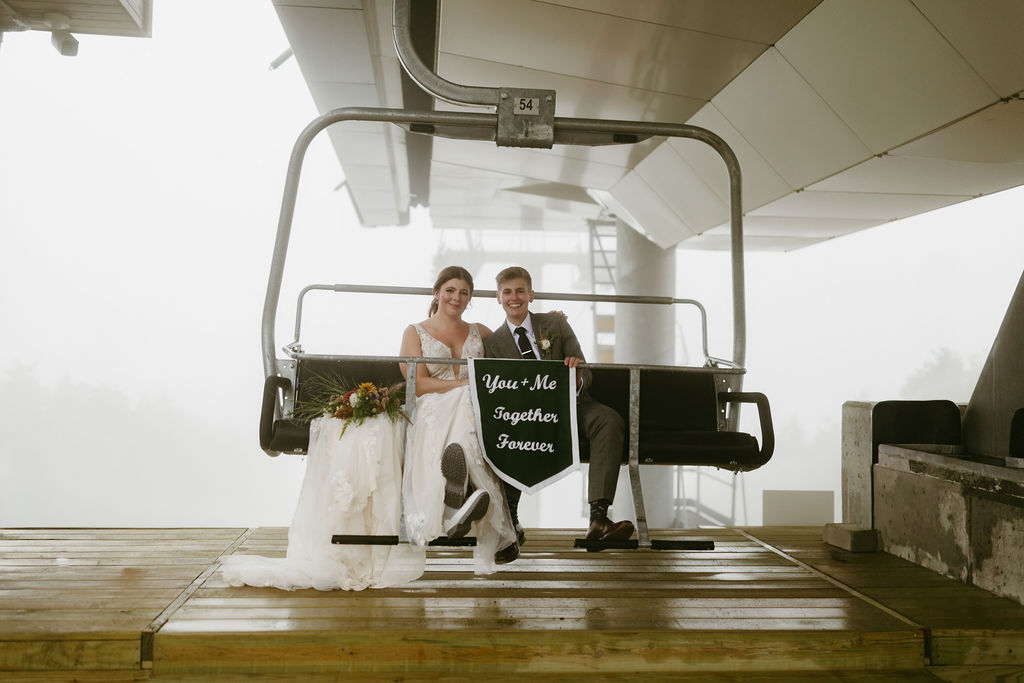 Brides, LGBTQ couple smiling at the camera with wedding banner that reads "you + me together forever" while sitting on a ski lift at Saddle Back Mountain in Rangeley Maine 