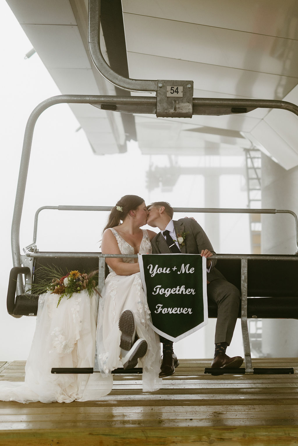 Brides, LGBTQ couple kissing with wedding banner that reads "you + me together forever" while sitting on a ski lift at Saddle Back Mountain in Rangeley Maine 